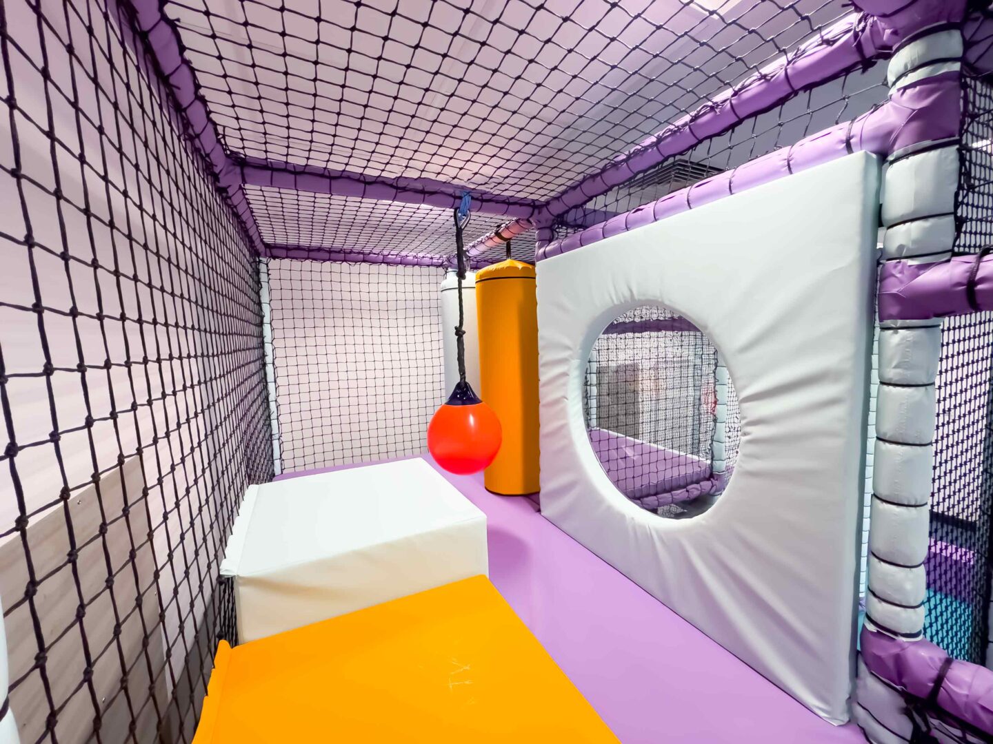Inside Trinity School's indoor softplay area. The theme of the softplay is orange, white and purple. There is an swinging buoy and a dome window.