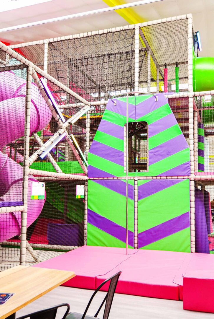 Fireman Pole with Matting at Fun Factory's indoor softplay area. There is a chevron wall which is purple and green that frames the fireman pole and the crashmats are pink.
