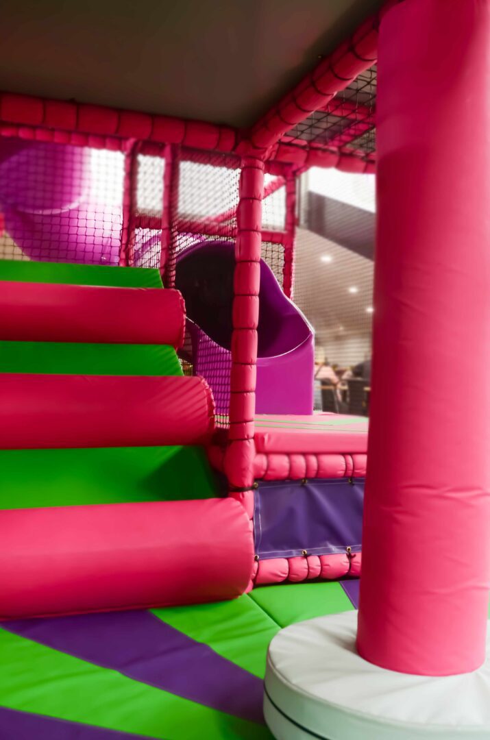 This is the bottom section of the toddler area, featuring a pink and white dizzy disc with green and purple applique flooring. This is found at the bottom of the purple tube slide. There is also a pink and green log ramp next to the slide leading to the next level.