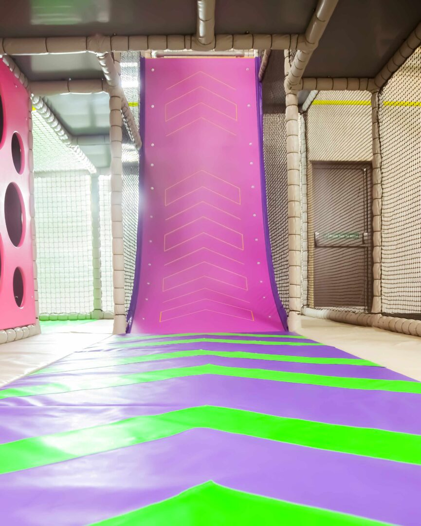 This is the Vertical Wall, a softplay equipment designed to be run up. The pink wall is matched with a green and purple chevron crashmat flooring.